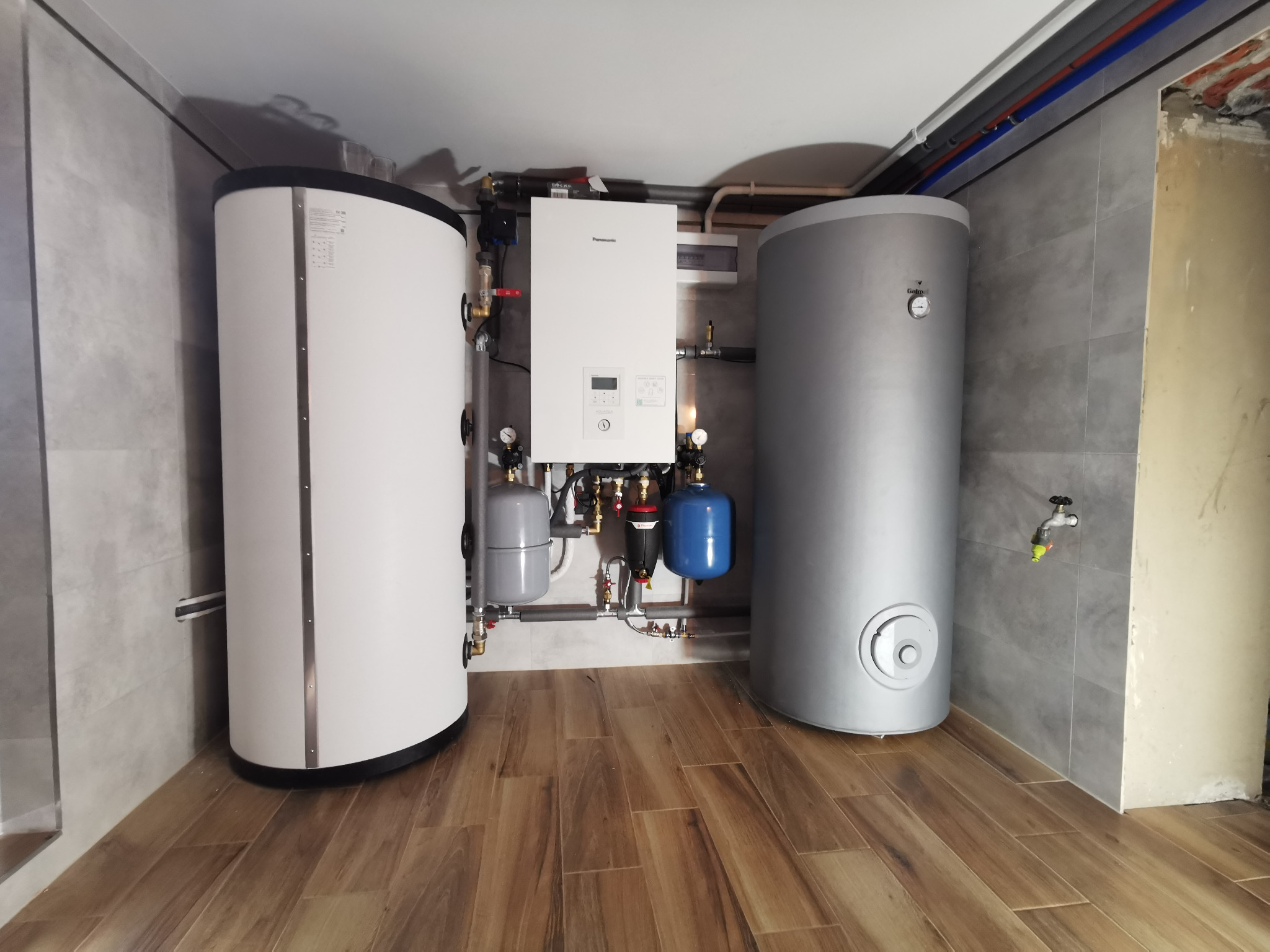 Kompletna instalacja grzewcza z dwoma zbiornikami i kotłem Panasonic na tle szarych płytek i drewnopodobnej podłogi, widok z przodu.