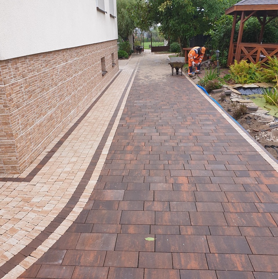 Ułożona ścieżka z kostki brukowej w kolorze brązowo-pomarańczowym, z obramowaniem z jasnej kostki, prowadząca wzdłuż ściany budynku z dekoracyjnym wykończeniem, w tle widoczny pracownik w stroju...