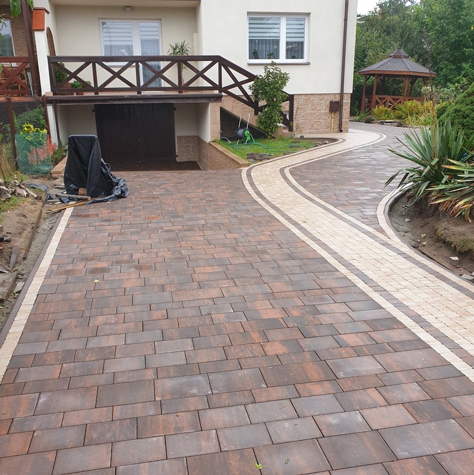 Nowo ułożony, brązowo-beżowy chodnik z kostki brukowej prowadzący do domu z drewnianą altaną w tle, widoczne ślady wilgoci na powierzchni.