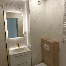 Nowoczesna łazienka z białymi płytkami imitującymi marmur z brązowymi żyłami, podświetlanym lustrem nad białą szafką z umywalką i złotą baterią, oraz toaletą z zabudowanym modułem spłukującym...