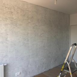 Remontownia Wnętrz - Ściana w pokoju po nałożeniu dekoracyjnej, szarej farby strukturalnej, widoczny grzejnik, drabina i kartony zabezpieczające podłogę.