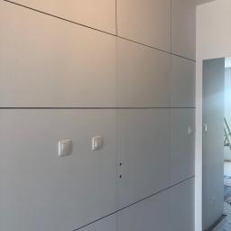 Remontownia Wnętrz - Szara zabudowa meblowa na wymiar z czarnymi liniami podziału, dwa włączniki światła i widoczne otwory na zamki, w tle fragment pokoju z drabiną.