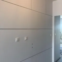 Szara zabudowa meblowa na wymiar z czarnymi liniami podziału, dwa włączniki światła i widoczne otwory na zamki, w tle fragment pokoju z drabiną.