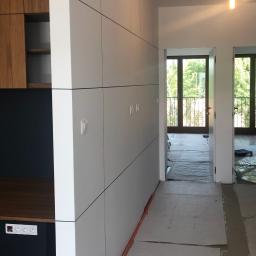 Remontownia Wnętrz - Wnętrze w trakcie prac wykończeniowych: biała ściana działowa z cienkimi czarnymi pasami, w tle widoczne drzwi i okna z widokiem na zieleń, podłoga zabezpieczona kartonem.