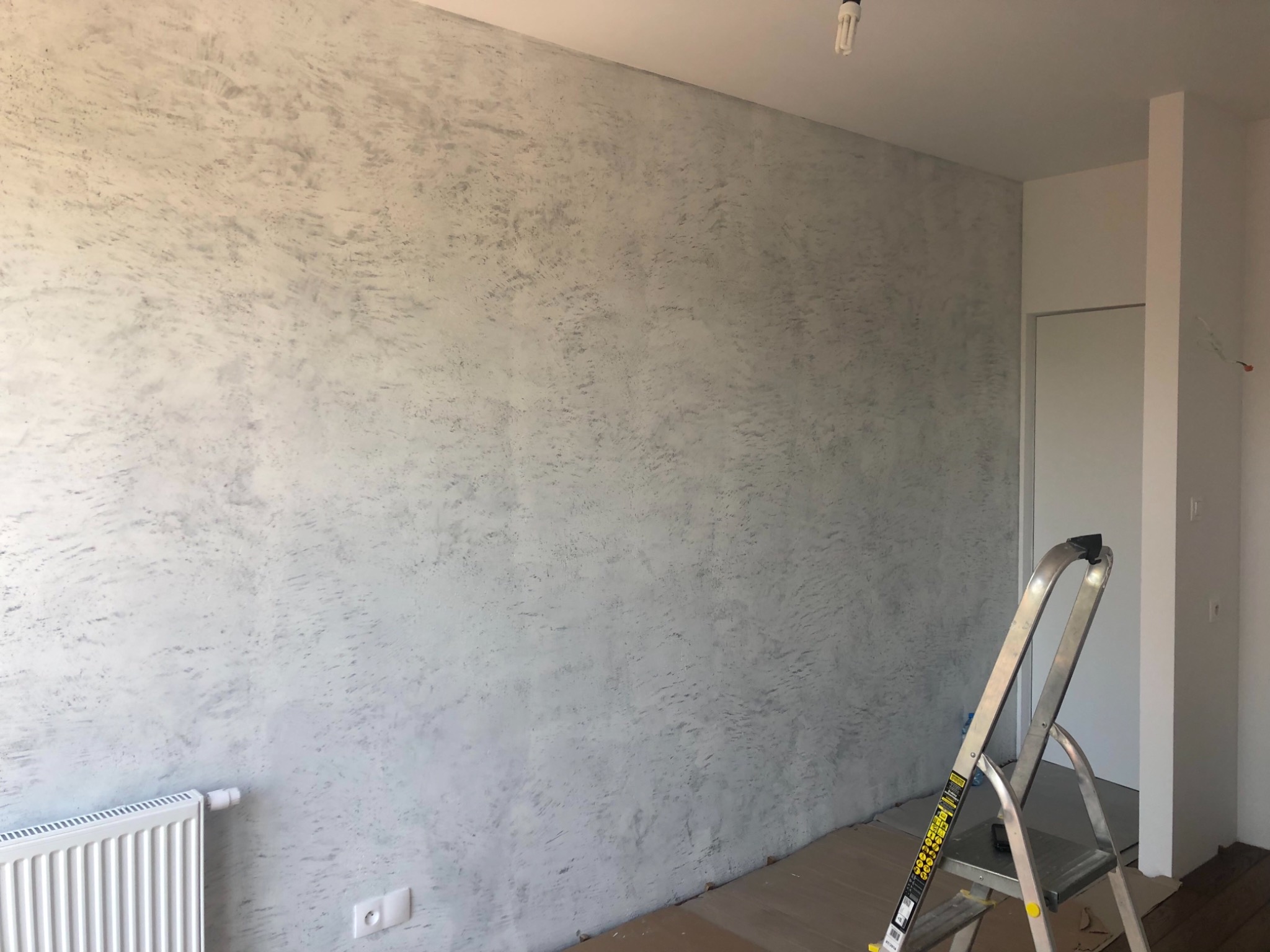 Ściana w pokoju po nałożeniu dekoracyjnej, szarej farby strukturalnej, widoczny grzejnik, drabina i kartony zabezpieczające podłogę.
