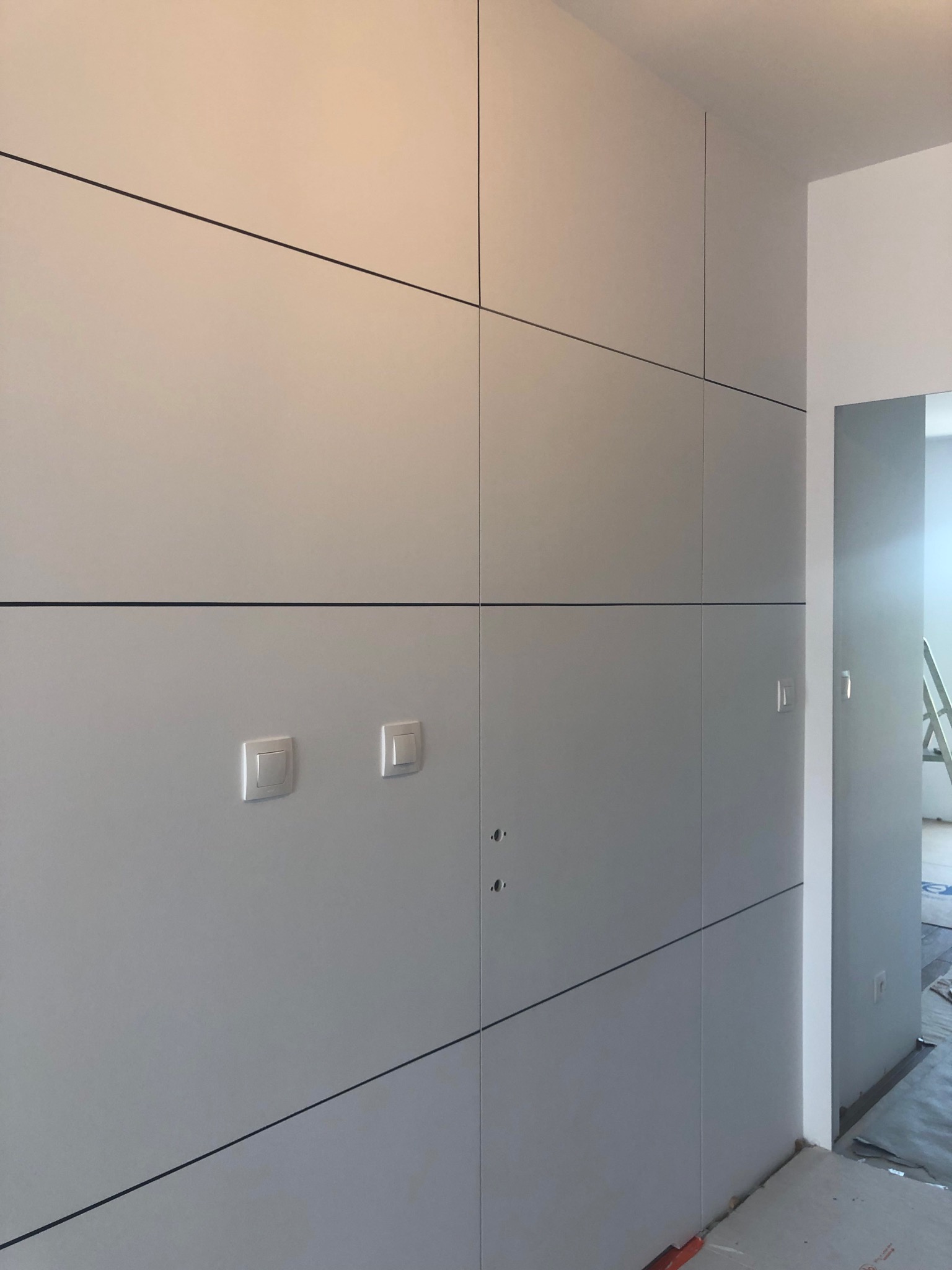 Szara zabudowa meblowa na wymiar z czarnymi liniami podziału, dwa włączniki światła i widoczne otwory na zamki, w tle fragment pokoju z drabiną.