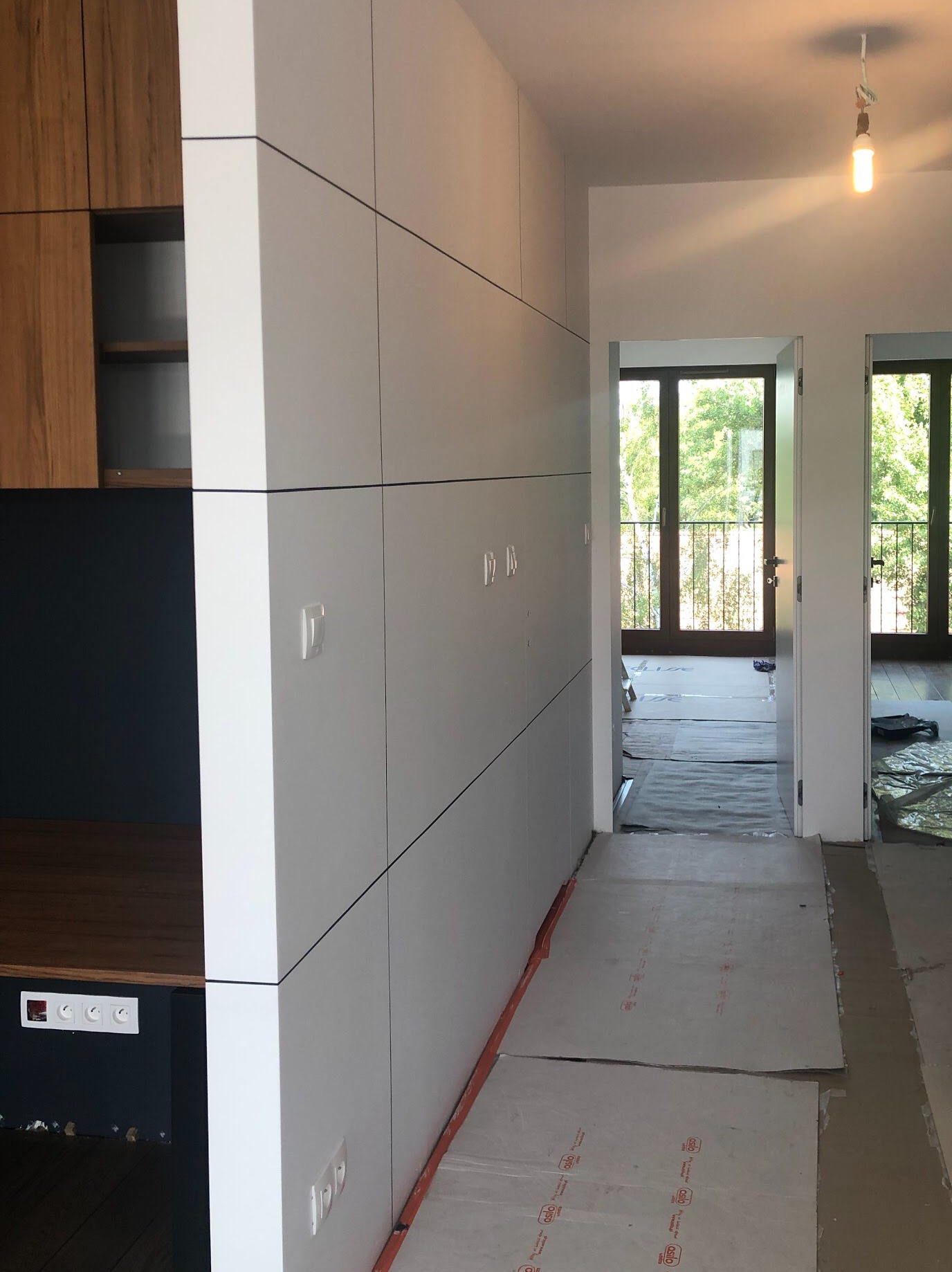 Wnętrze w trakcie prac wykończeniowych: biała ściana działowa z cienkimi czarnymi pasami, w tle widoczne drzwi i okna z widokiem na zieleń, podłoga zabezpieczona kartonem.