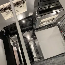 Wnętrze biura w trakcie przygotowań do malowania: zabezpieczona folią podłoga, białe ściany i czarne kolumny, aluminiowa drabina oparta o ścianę, oświetlenie punktowe i dekoracyjne panele sufitowe...