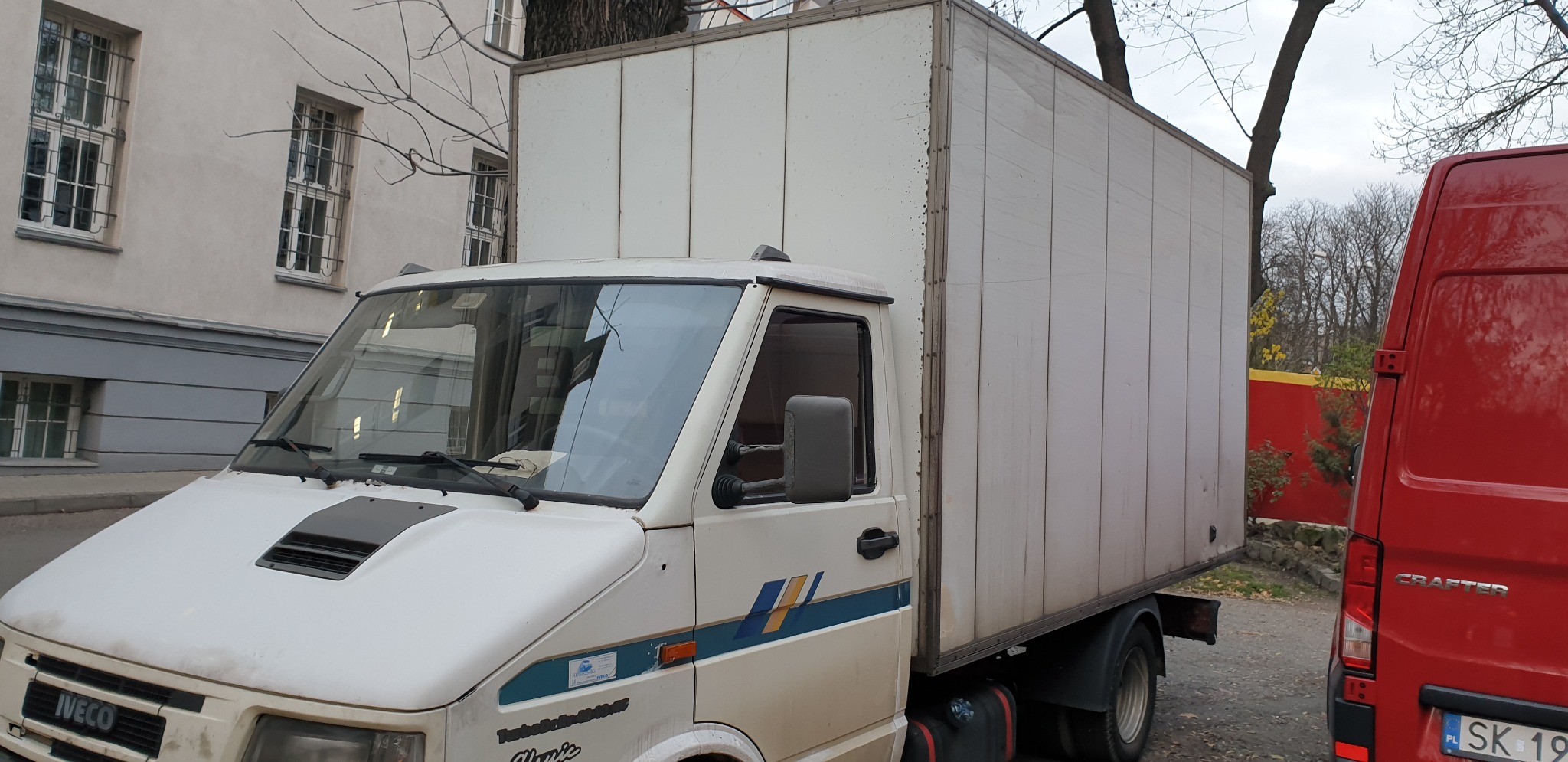 Biały, lekko zabrudzony samochód dostawczy Iveco z kontenerem na tle budynku z oknami z kratami i czerwonego samochodu dostawczego.