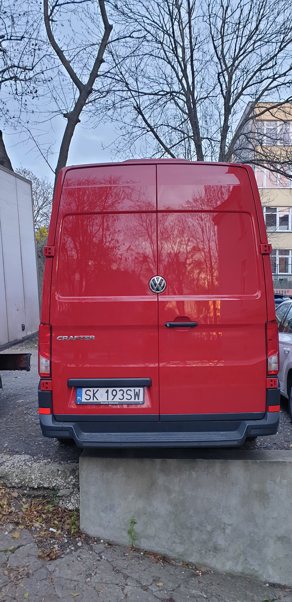 Tył czerwonego busa Volkswagen Crafter z tablicą rejestracyjną SK 193SW na tle drzew bez liści i budynku.