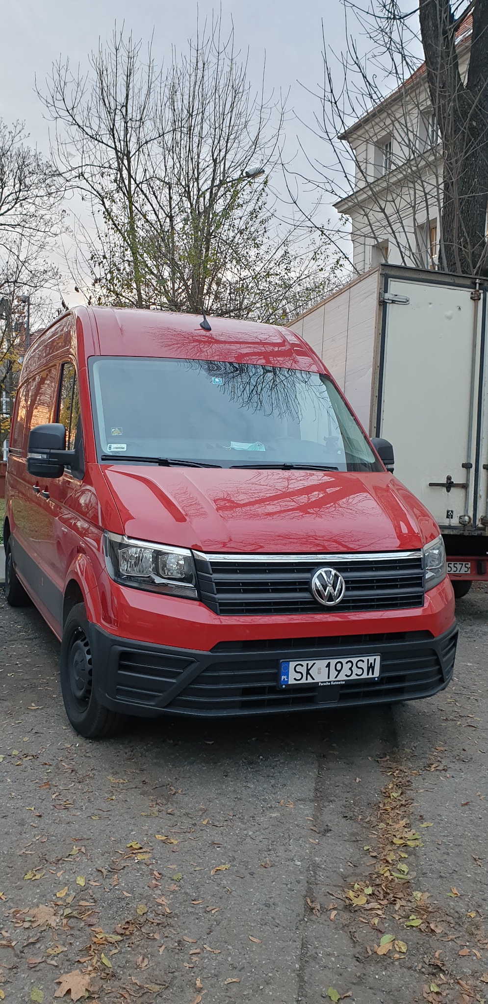 Czerwony bus Volkswagen Crafter zaparkowany na zewnątrz, widoczny z przodu, z tablicą rejestracyjną SK 193SW, obok zaparkowana biała przyczepa transportowa, jesienne drzewa w tle.