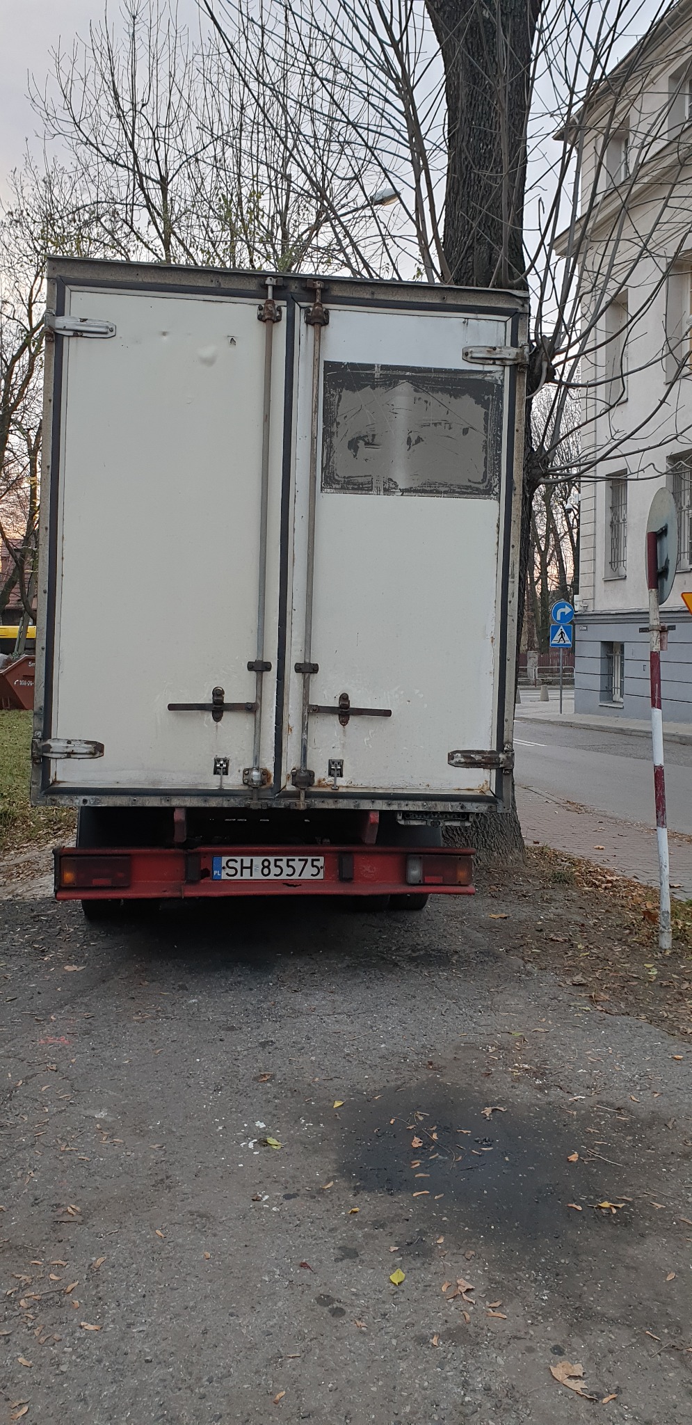 Tył białego, lekko uszkodzonego busa z widoczną tablicą rejestracyjną SH 85575, zaparkowanego przy chodniku obok drzewa w jesiennej scenerii miejskiej.