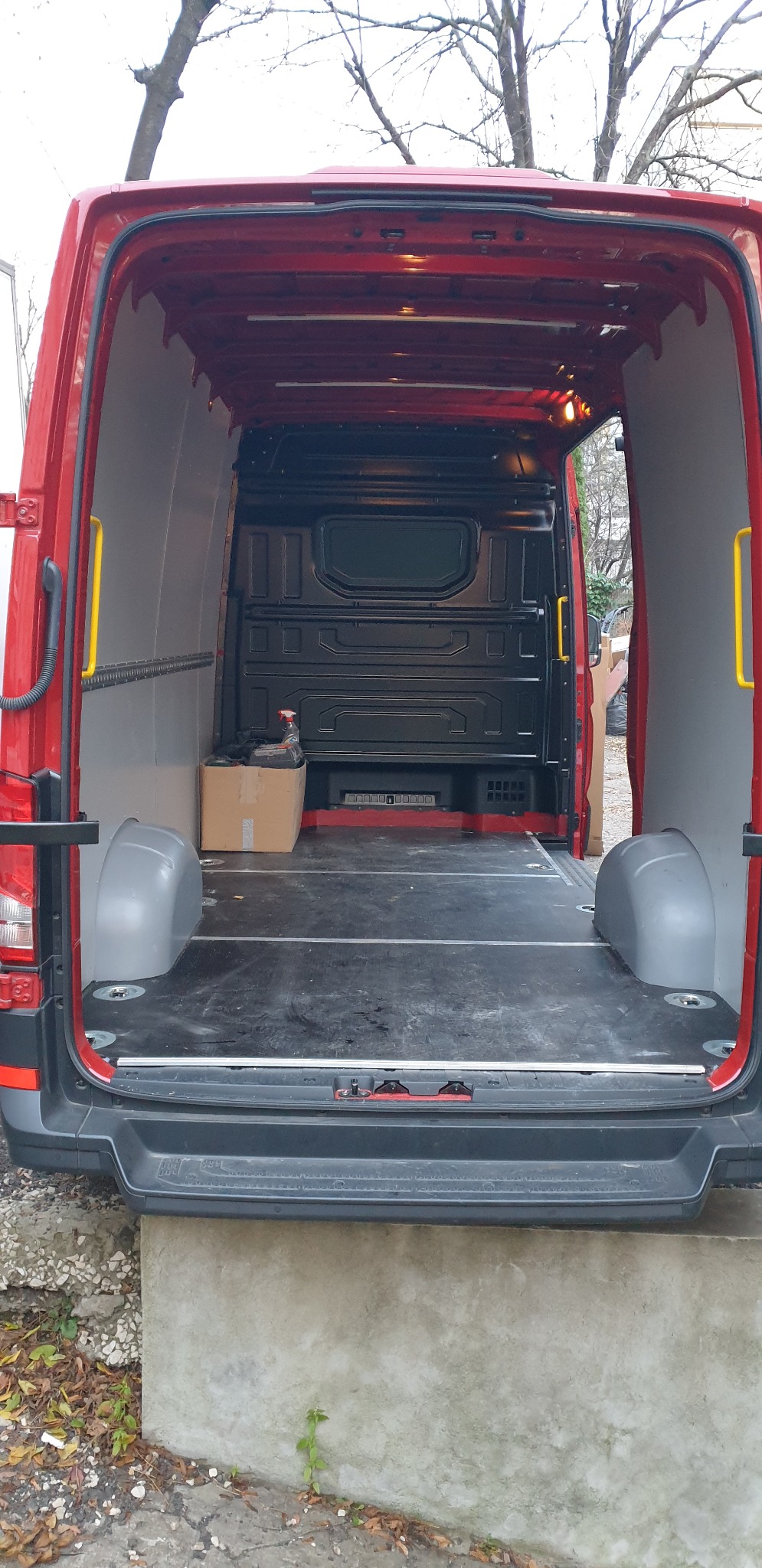 Wnętrze pustej, czerwonej furgonetki z zabezpieczonymi ścianami i podłogą, widok z tyłu na betonową rampę załadunkową i karton z płynem do czyszczenia.