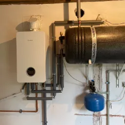 Zainstalowany gazowy kocioł kondensacyjny Bosch z zasobnikiem Kospel, rury w szarej izolacji, nieotynkowana ściana.