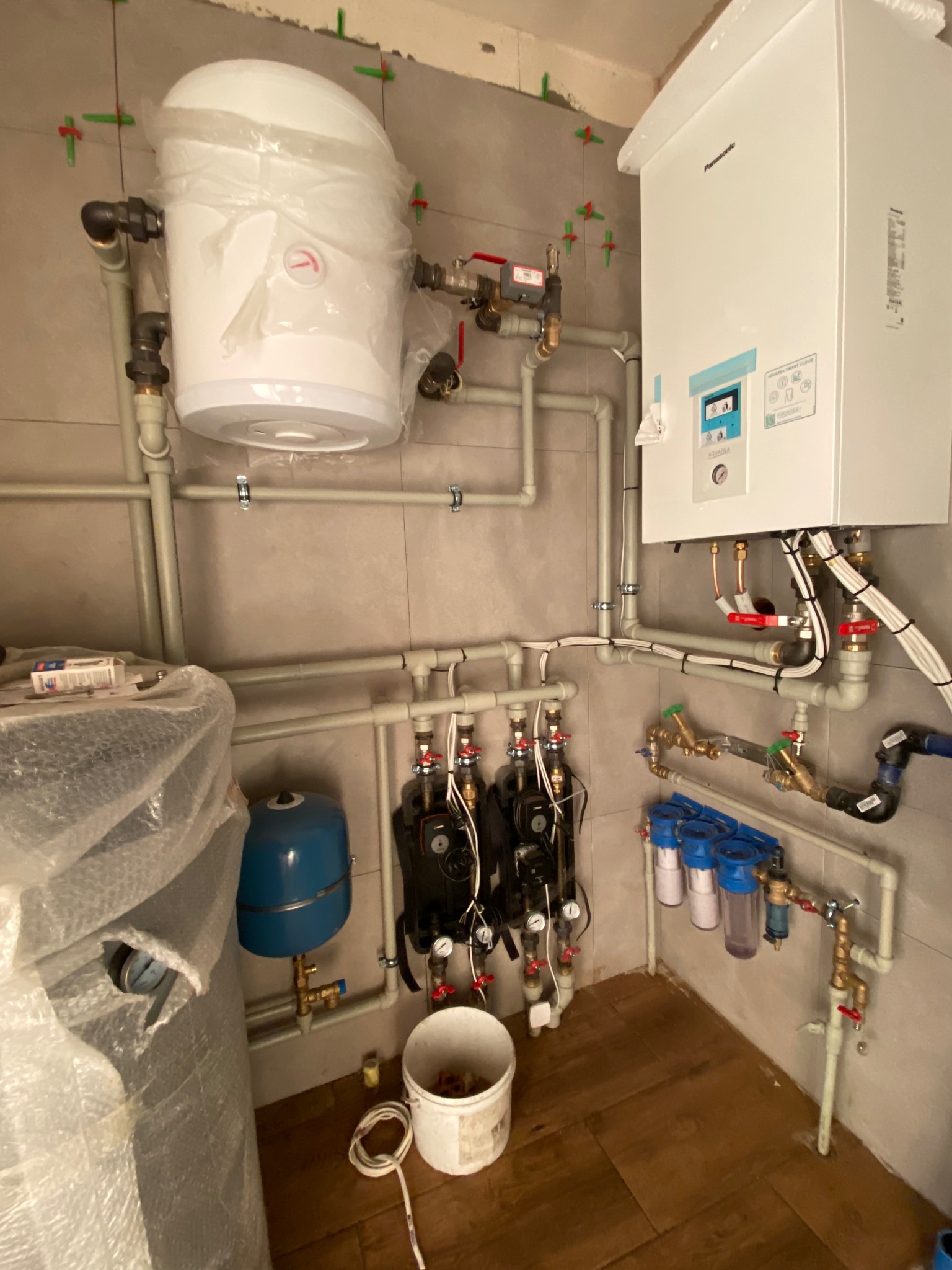 Złożona instalacja grzewcza z podgrzewaczem wody w folii ochronnej, pompami obiegowymi, filtrami i kotłem gazowym Panasonic, zamontowana na ścianie wyłożonej płytkami ceramicznymi, widoczne...
