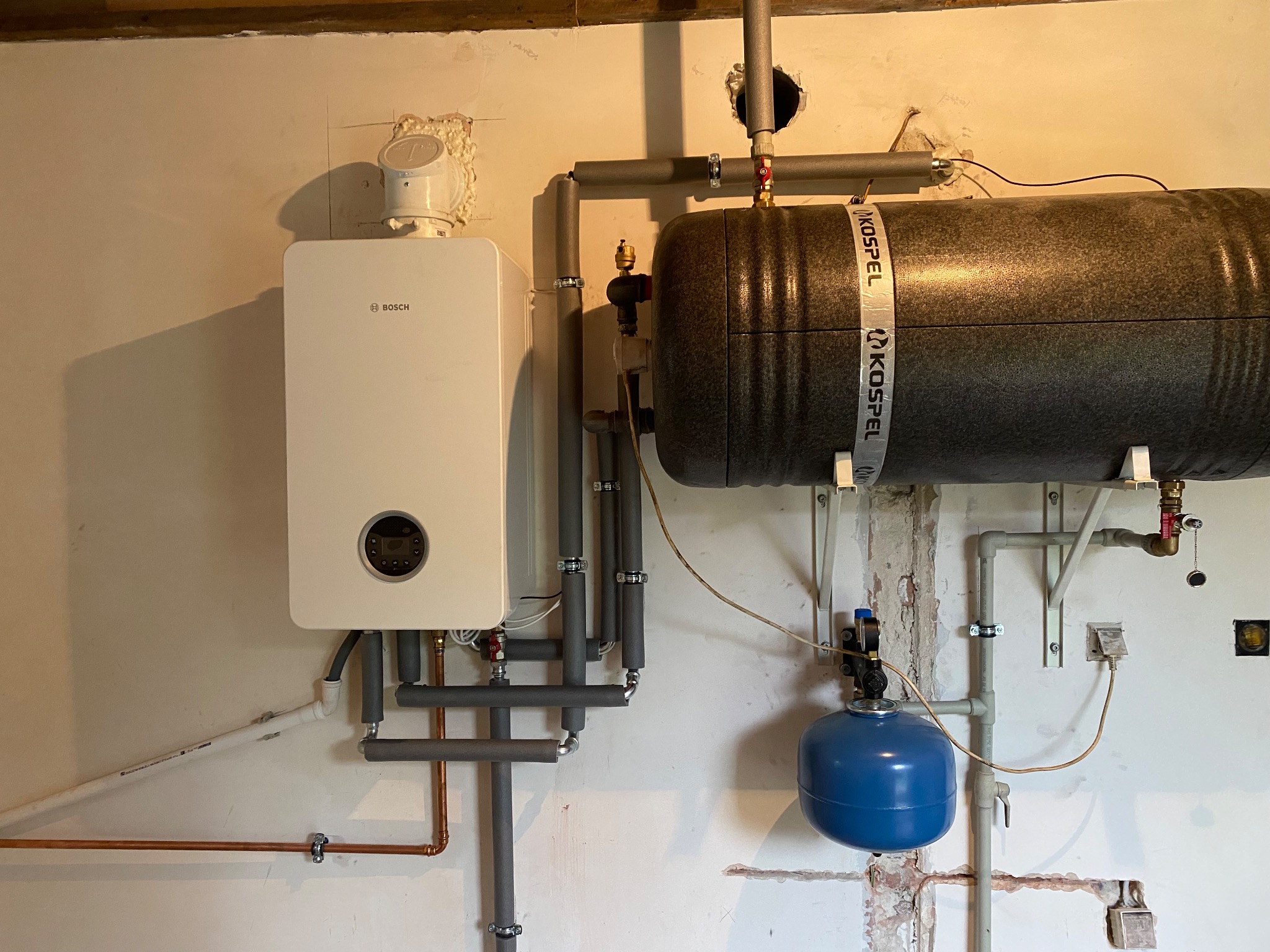 Zainstalowany gazowy kocioł kondensacyjny Bosch z zasobnikiem Kospel, rury w szarej izolacji, nieotynkowana ściana.