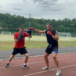 Dwóch mężczyzn w rękawicach bokserskich trenuje na bieżni lekkoatletycznej, jeden w czarnej koszulce bez rękawów wyprowadza cios, drugi w czerwonej koszulce przyjmuje pozycję obronną, w tle drzewa...