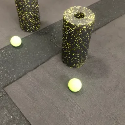 Dwa czarne, teksturowane wałki do masażu z żółtymi plamkami i dwie zielone piłeczki tenisowe na szarej macie do ćwiczeń.