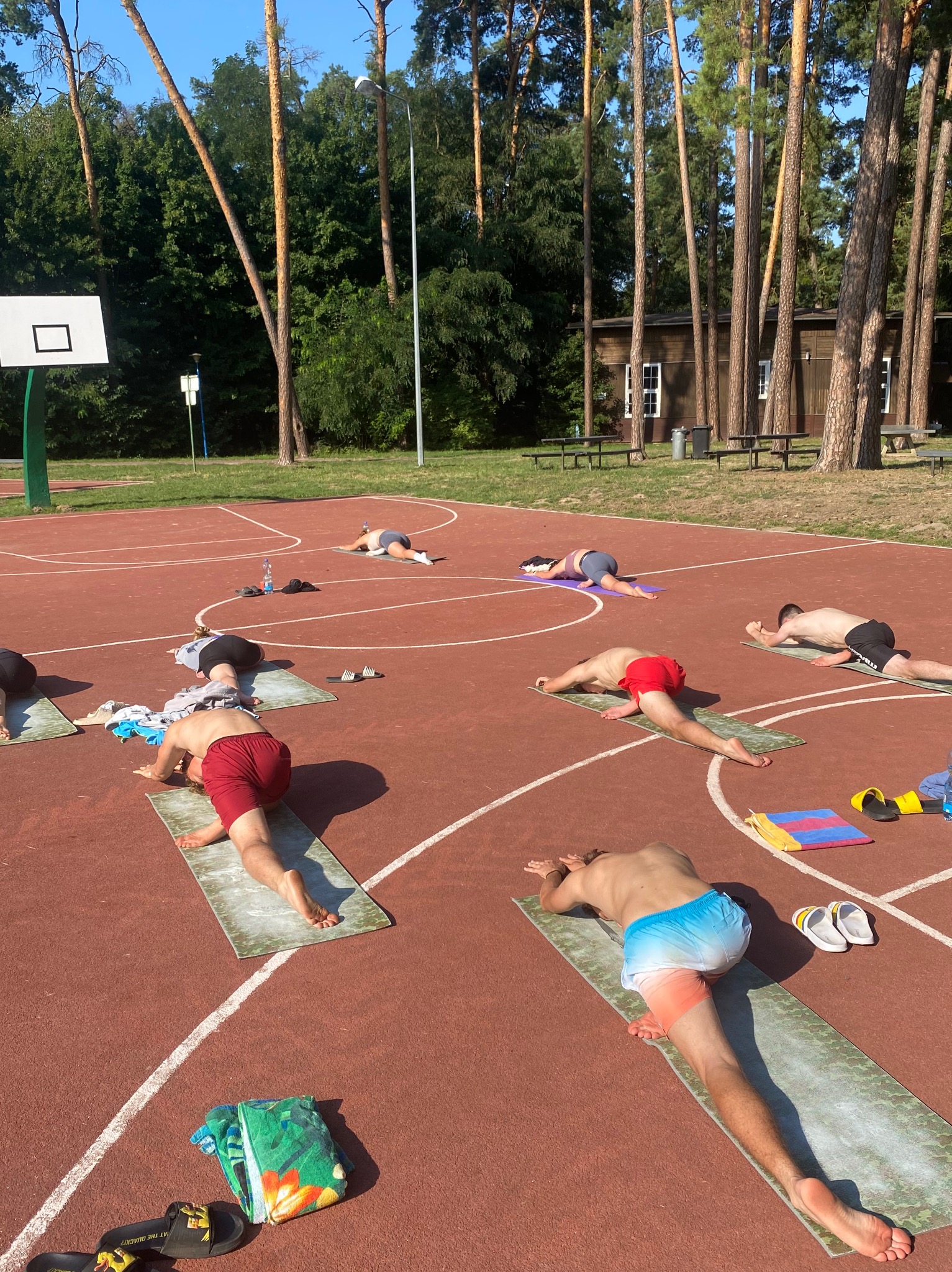 Grupa osób ćwiczy na matach do jogi na czerwonym boisku do koszykówki w otoczeniu drzew i budynku, w słoneczny dzień.