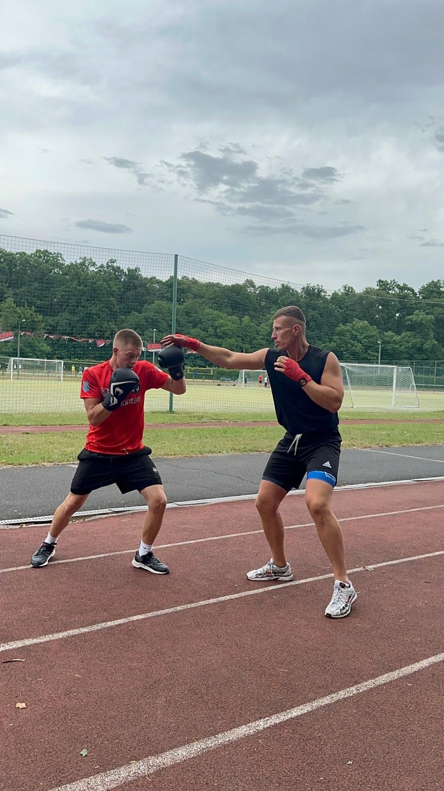 Dwóch mężczyzn w rękawicach bokserskich trenuje na bieżni lekkoatletycznej, jeden w czarnej koszulce bez rękawów wyprowadza cios, drugi w czerwonej koszulce przyjmuje pozycję obronną, w tle drzewa...