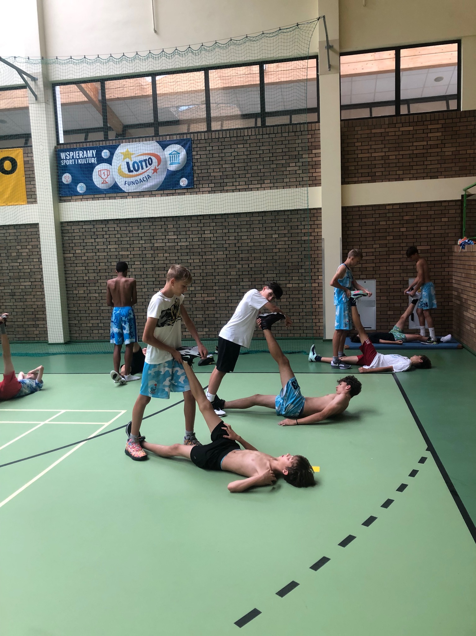 Grupa nastolatków w sali gimnastycznej wykonuje ćwiczenia rozciągające, jeden pomaga drugiemu podnosząc nogę, widoczny baner Lotto na ścianie.