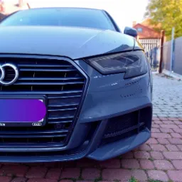 Szary samochód marki Audi, widok z przodu na grill z logo i reflektor, zaparkowany na czerwonej kostce brukowej przed metalowym ogrodzeniem.