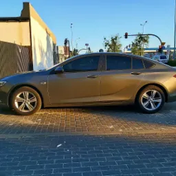 Szaro-złoty sedan zaparkowany na chodniku z kostki brukowej w słoneczny dzień, z widocznymi przyciemnionymi szybami i elementami otoczenia miejskiego.