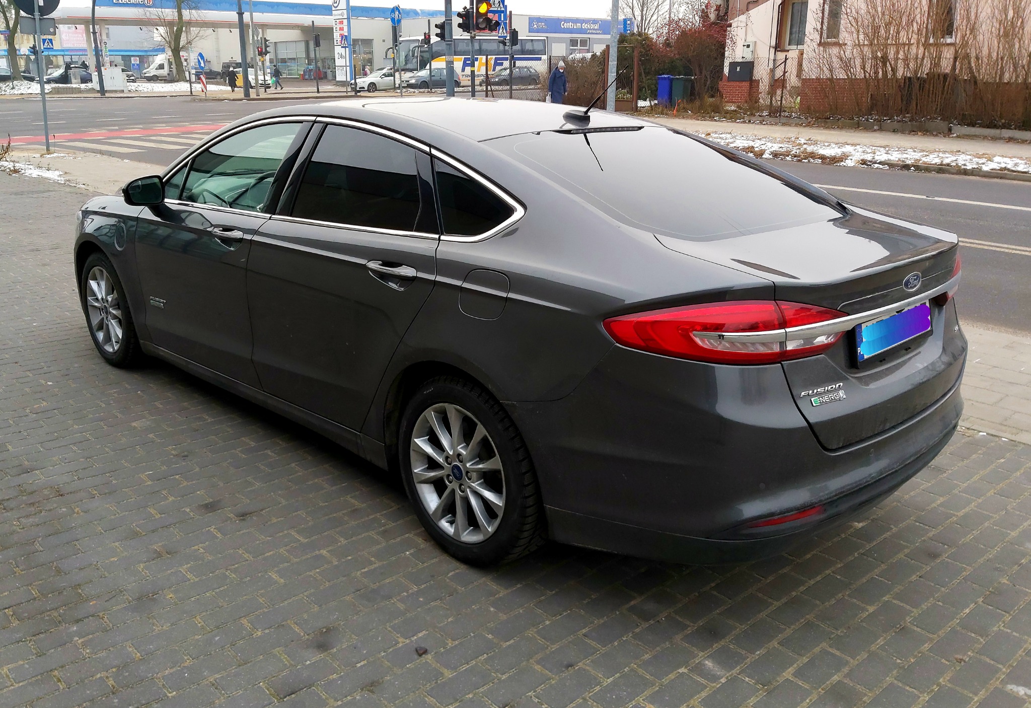 Ciemnoszary Ford Fusion z przyciemnionymi szybami, zaparkowany na brukowanej ulicy, widok z tyłu i boku, z widocznymi detalami jak logo Forda i oznaczenie 'Fusion Energi'.