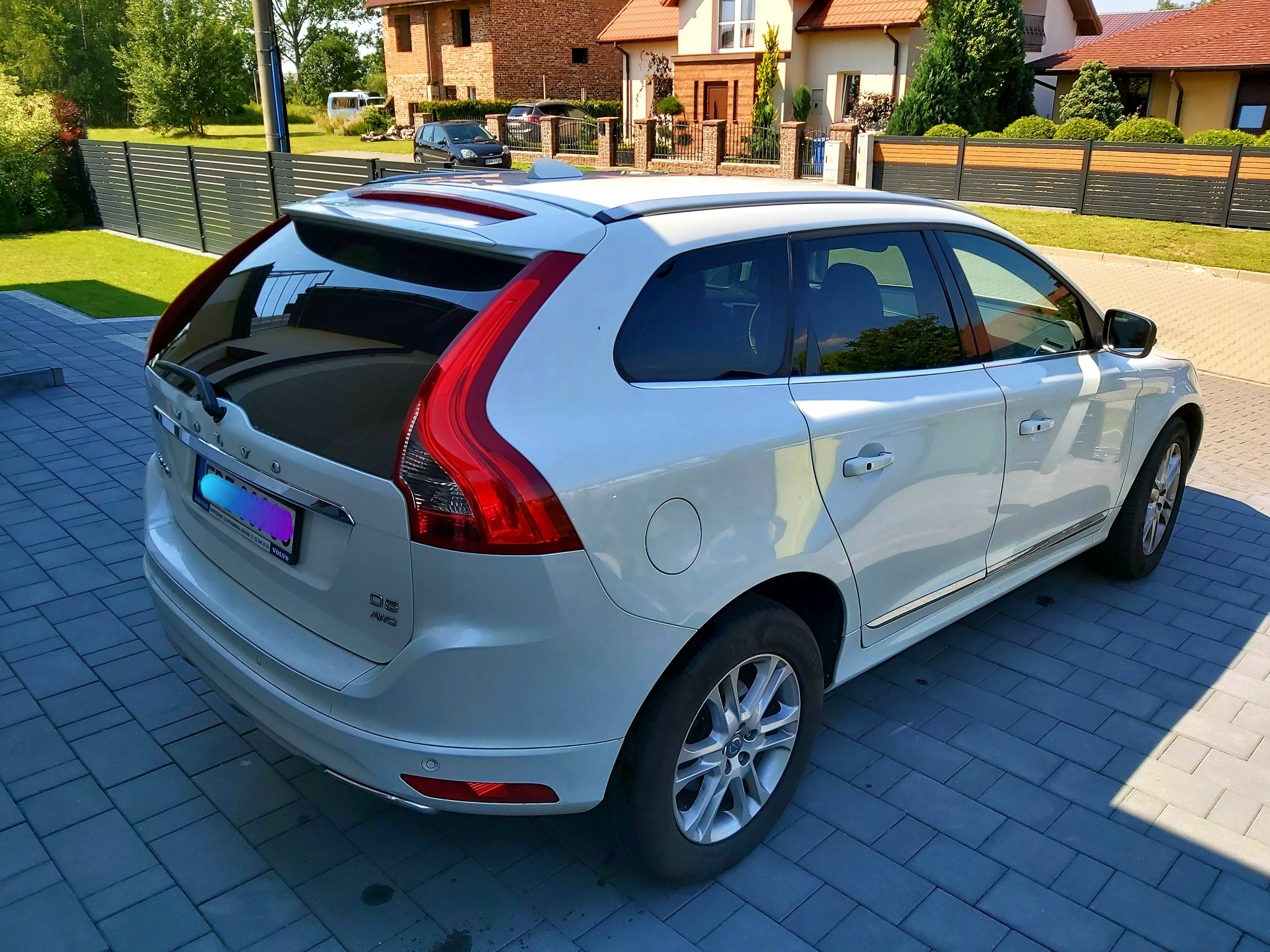 Białe Volvo XC60 zaparkowane na brukowanej nawierzchni przed domem jednorodzinnym, widok z tyłu, z widocznym logo i oznaczeniem modelu.