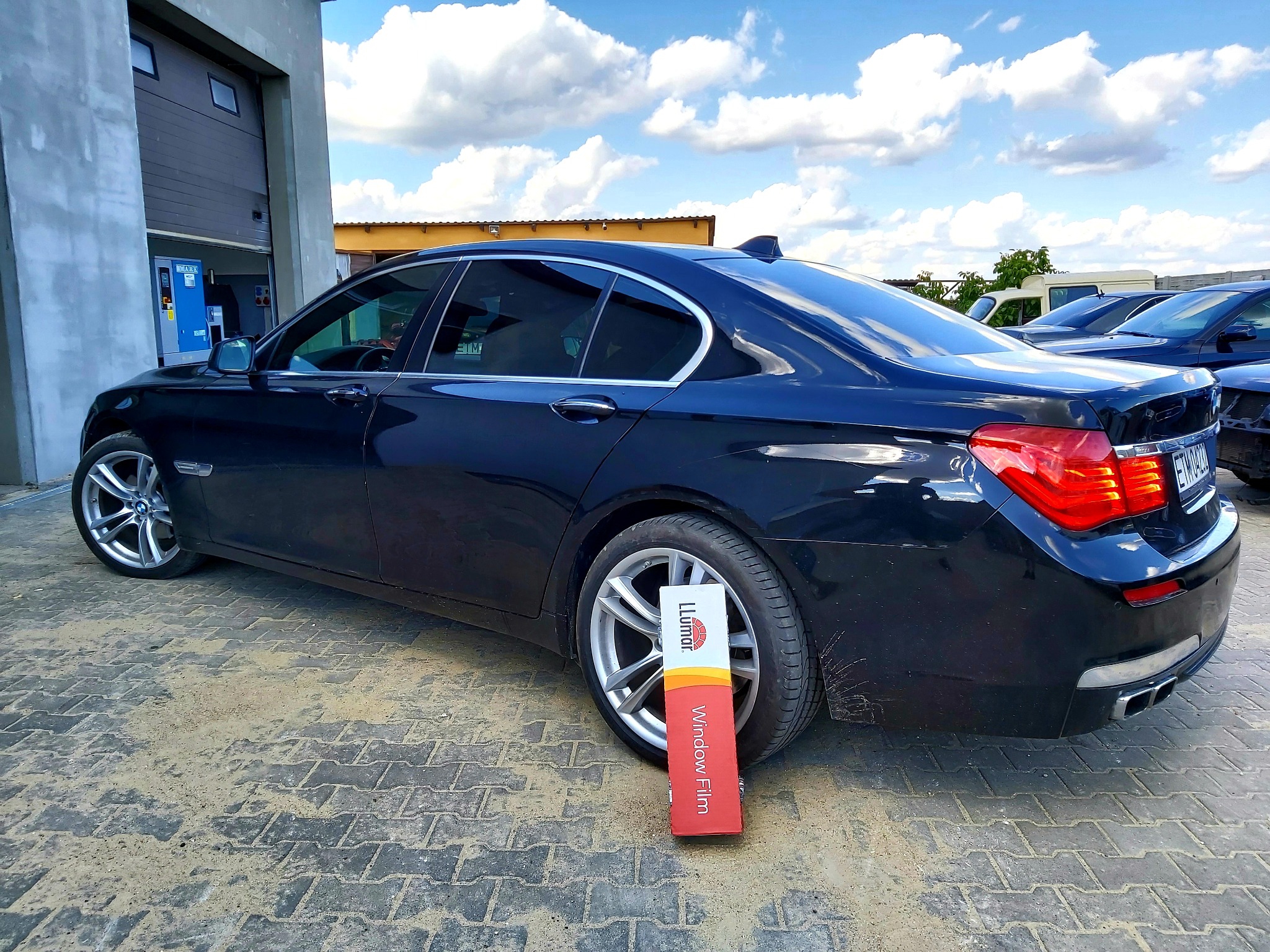 Ciemny sedan marki BMW, zaparkowany na kostce brukowej, z widoczną paczką folii okiennej marki Llumar opartą o tylne koło, sugerującą proces przyciemniania szyb.