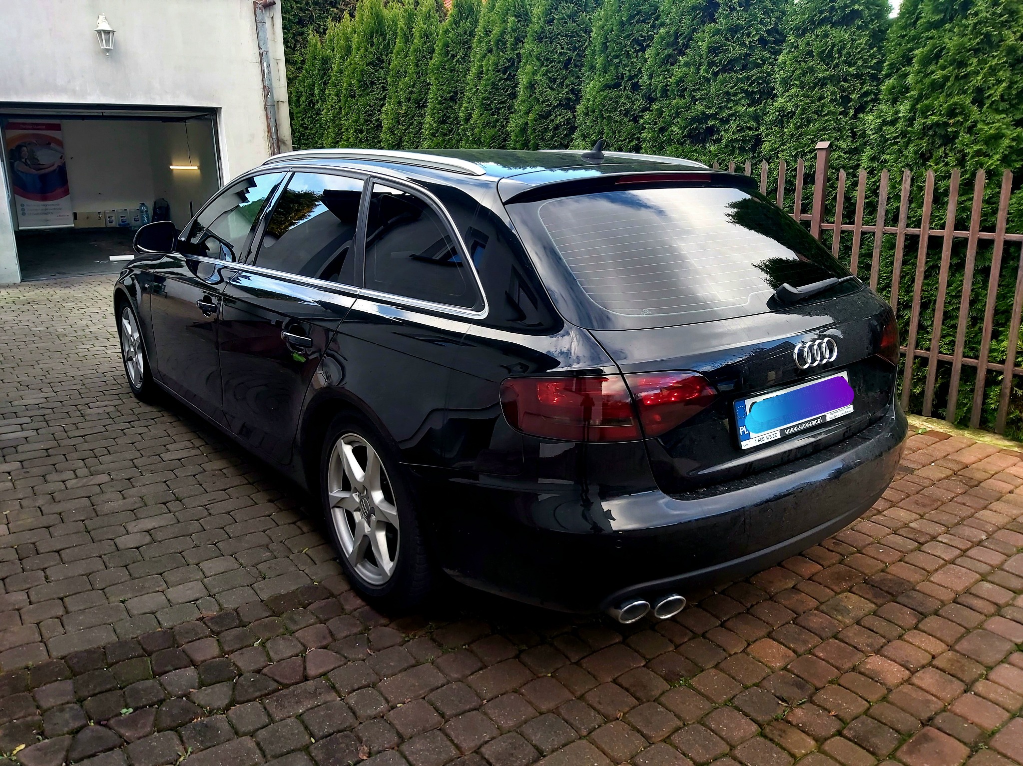 Czarny samochód typu kombi marki Audi z przyciemnionymi tylnymi szybami, zaparkowany na kostce brukowej przed garażem i żywopłotem.