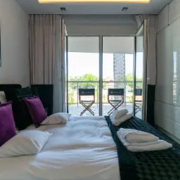 Nowoczesna sypialnia z balkonem: biała pościel, fioletowe poduszki, czarna narzuta, widok na zewnątrz z dwoma krzesłami reżyserskimi.