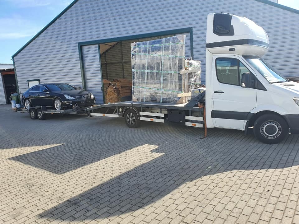 Czarny Mercedes CLS na lawecie za białym samochodem dostawczym z kabiną sypialną, przewożącym zabezpieczony ładunek, na tle magazynu z otwartą bramą.