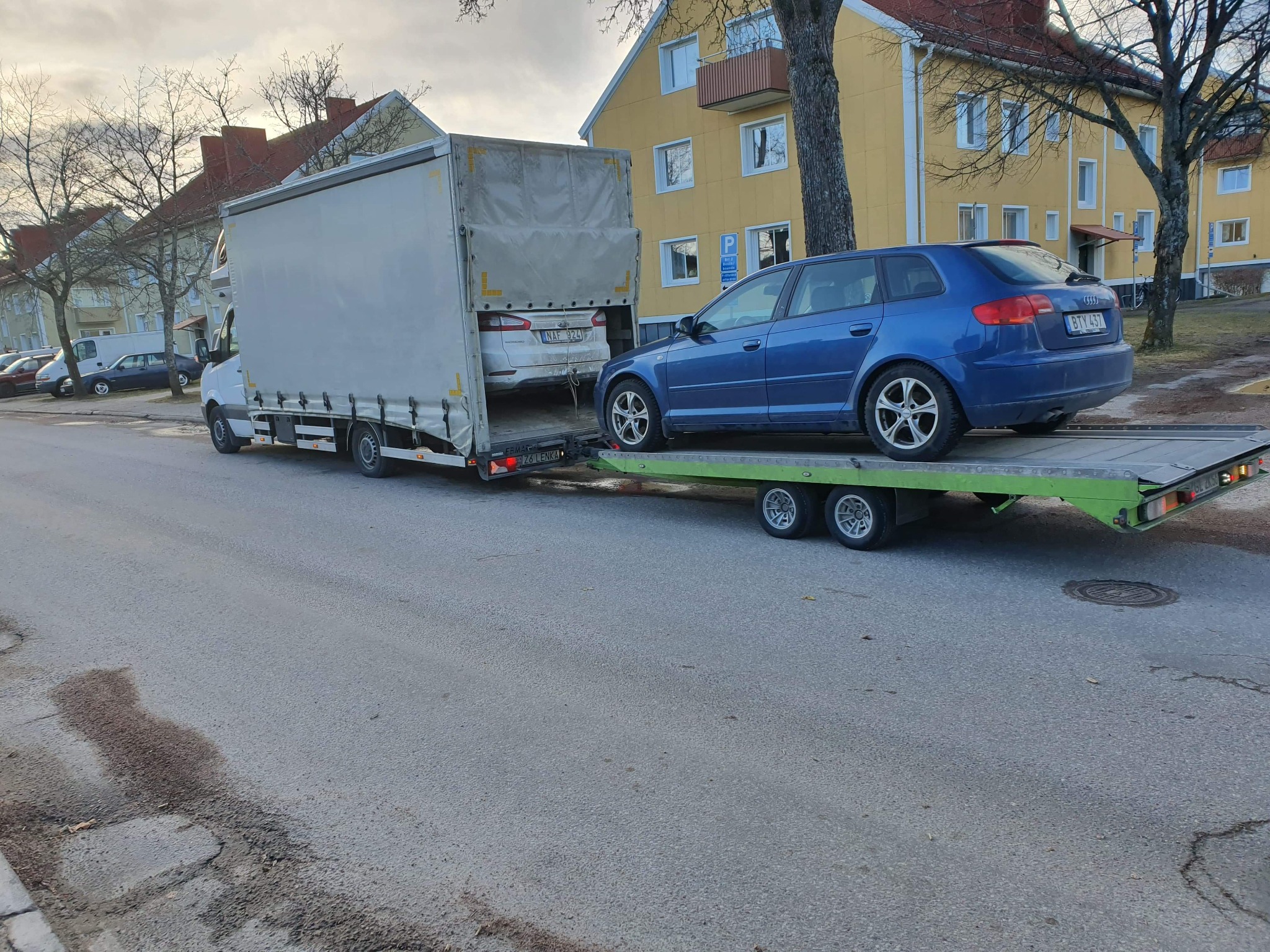 Ciężarówka z przyczepą lawetą przewożąca dwa samochody osobowe na tle zabudowy miejskiej.