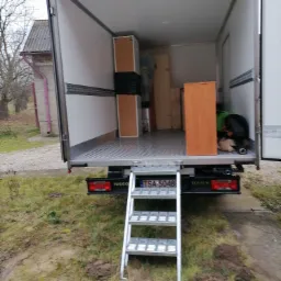 Wnętrze otwartego busa transportowego z rampą załadowczą, częściowo załadowane meblami, widok z tyłu na zewnątrz na trawnik i budynek gospodarczy.