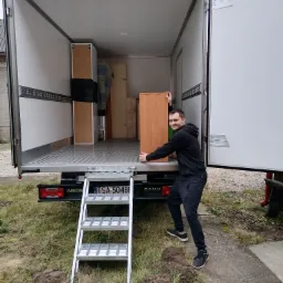 Mężczyzna w czarnym dresie wkłada drewnianą szafkę do wnętrza białej furgonetki z otwartymi drzwiami, widoczne schodki wejściowe i częściowo załadowane meble w środku.