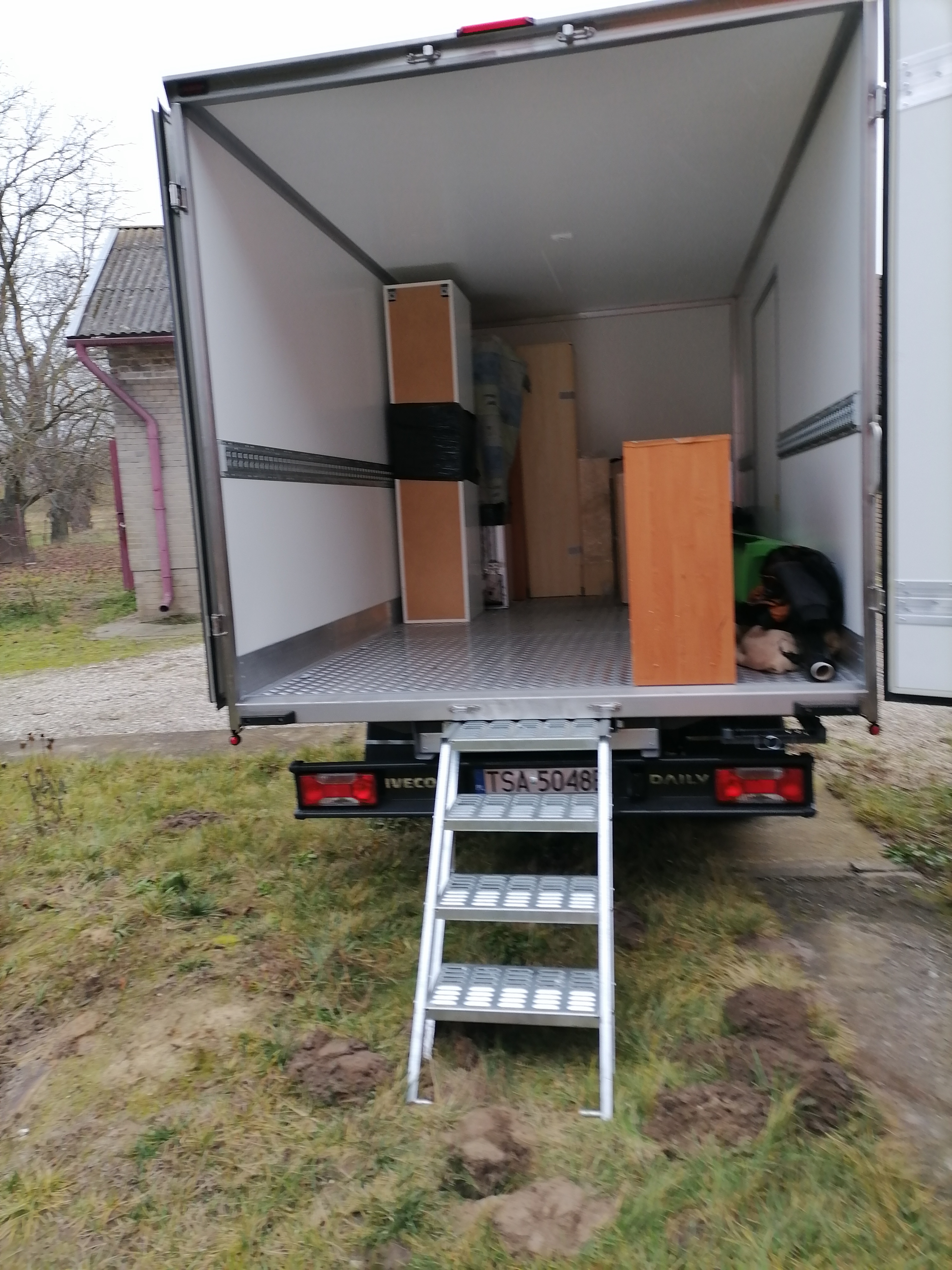 Wnętrze otwartego busa transportowego z rampą załadowczą, częściowo załadowane meblami, widok z tyłu na zewnątrz na trawnik i budynek gospodarczy.