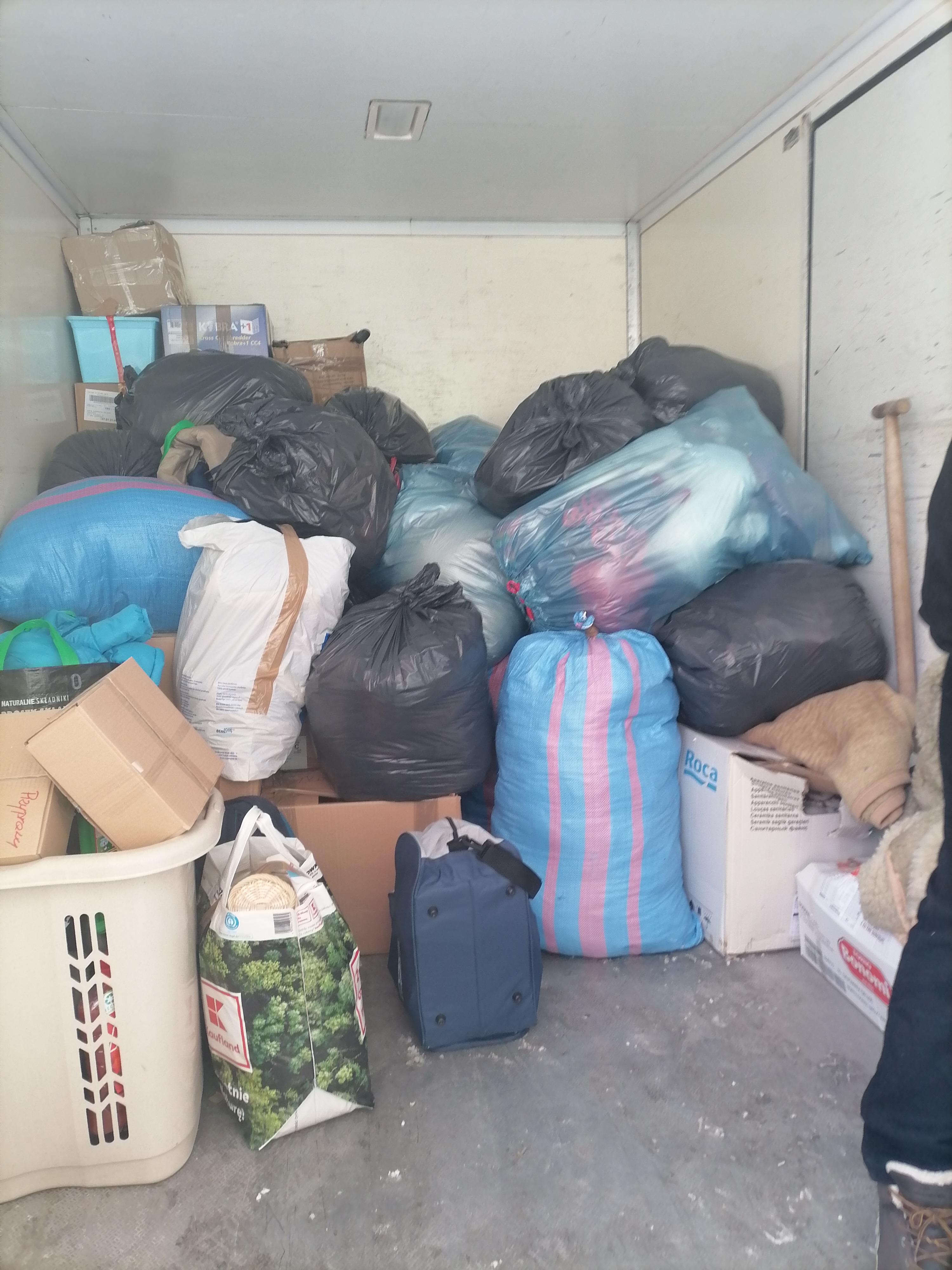 Wnętrze furgonetki wypełnionej workami na śmieci, kartonami i torbami, gotowe do transportu. Widoczna torba z logiem Kaufland oraz pudełko Roca.