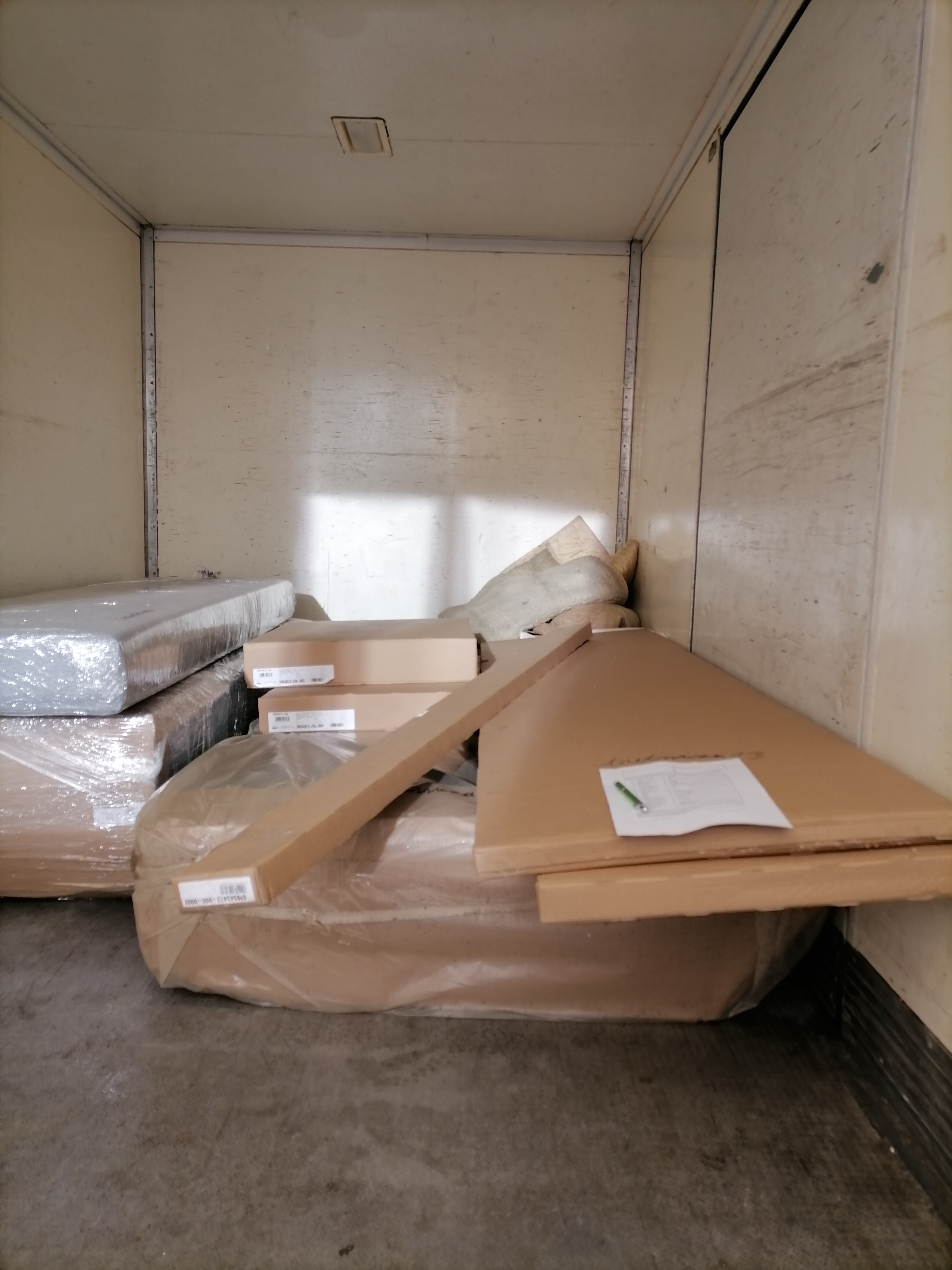 Wnętrze skrzyni ładunkowej z zapakowanymi meblami w folii i kartonach, widoczne etykiety adresowe i zielony długopis na wierzchu paczki.