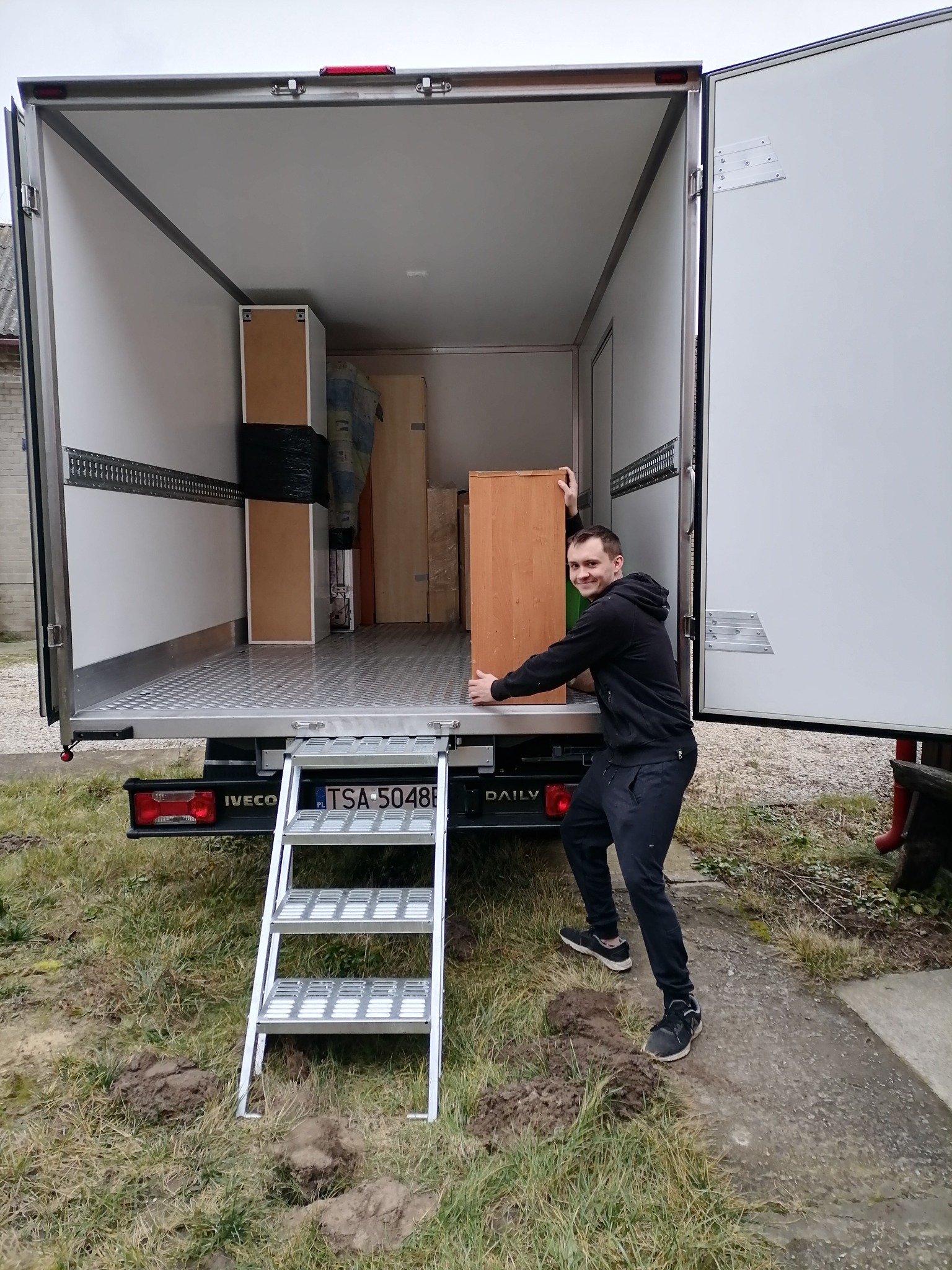 Mężczyzna w czarnym dresie wkłada drewnianą szafkę do wnętrza białej furgonetki z otwartymi drzwiami, widoczne schodki wejściowe i częściowo załadowane meble w środku.