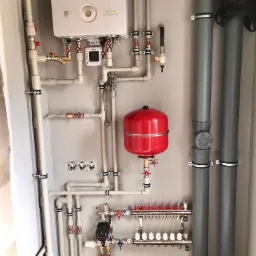 Złożony system rur, zaworów i urządzeń grzewczych na szarej ścianie, w tym biały piec, czerwony zbiornik wyrównawczy i rozdzielacz z białymi odpowietrznikami.