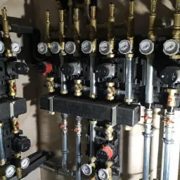 Rozdzielacz instalacji grzewczej z pompami obiegowymi, manometrami i zaworami, widok z bliska na skomplikowany system rur i połączeń.