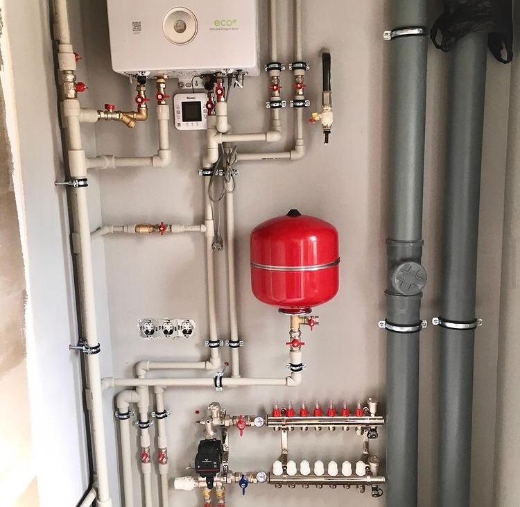 Złożony system rur, zaworów i urządzeń grzewczych na szarej ścianie, w tym biały piec, czerwony zbiornik wyrównawczy i rozdzielacz z białymi odpowietrznikami.