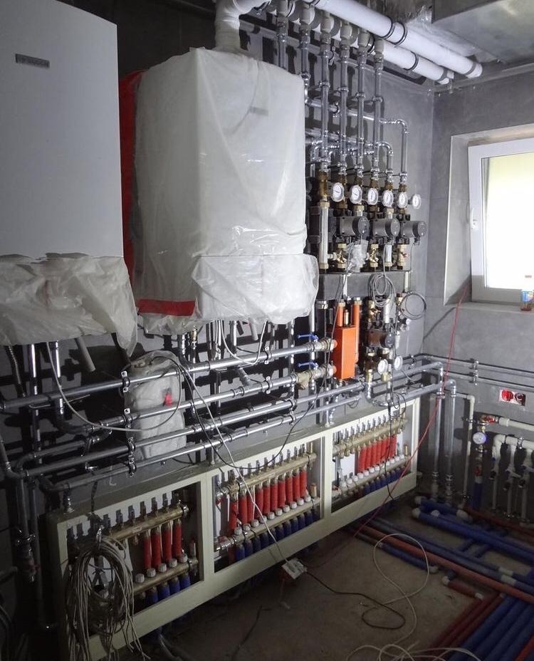 Rozbudowana instalacja grzewcza z kotłem i rozdzielaczem podłogówki, częściowo zabezpieczona folią ochronną, widoczne liczne rury, zawory i manometry w surowym wnętrzu.