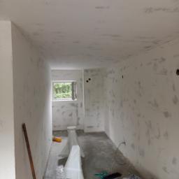 Krzepicki Piotr - Wnętrze pokoju w trakcie prac wykończeniowych: ściany i sufit pokryte szpachlą, widoczne narzędzia i wiadro z materiałem, naturalne światło przez okno.