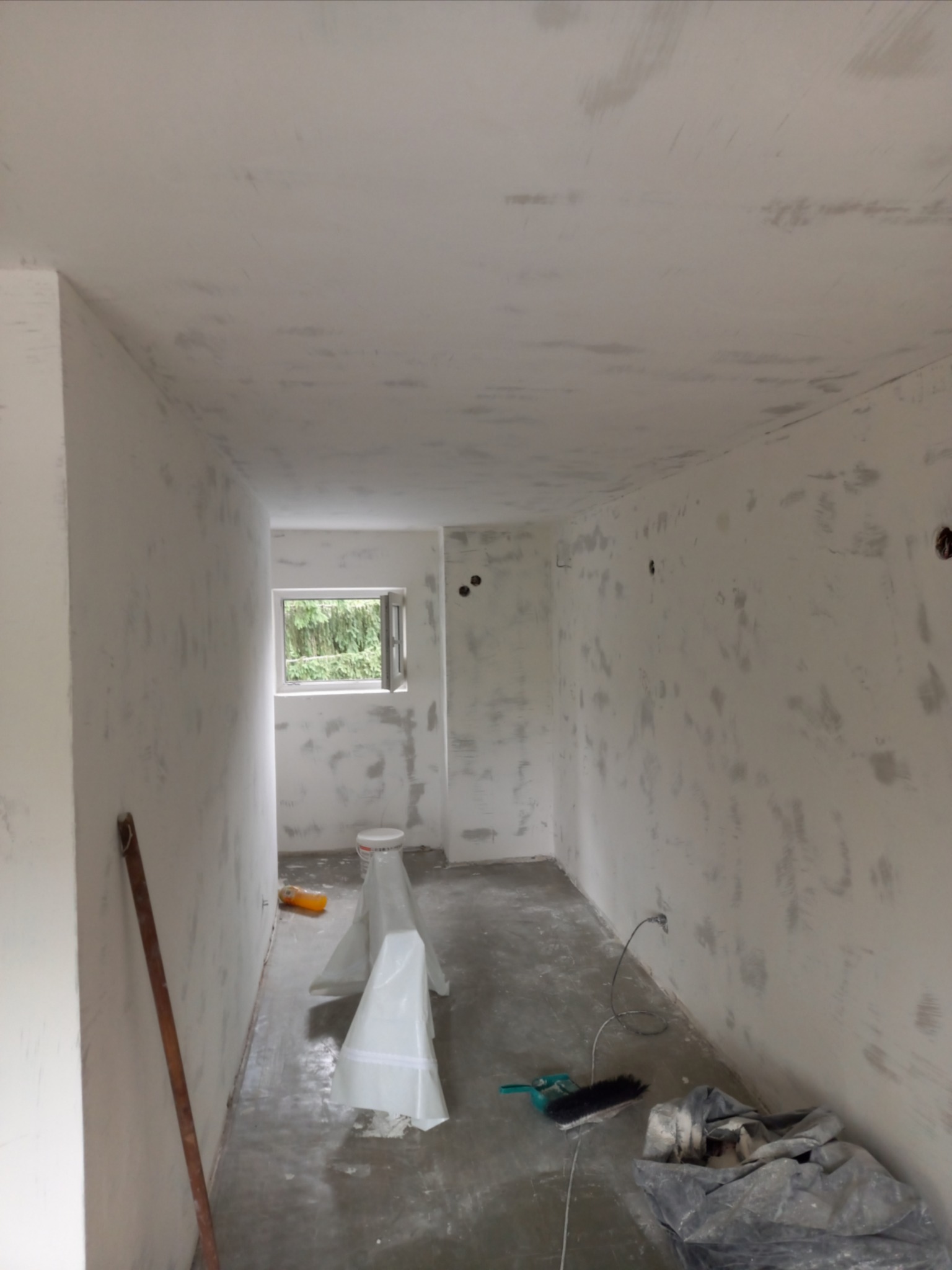 Wnętrze pokoju w trakcie prac wykończeniowych: ściany i sufit pokryte szpachlą, widoczne narzędzia i wiadro z materiałem, naturalne światło przez okno.