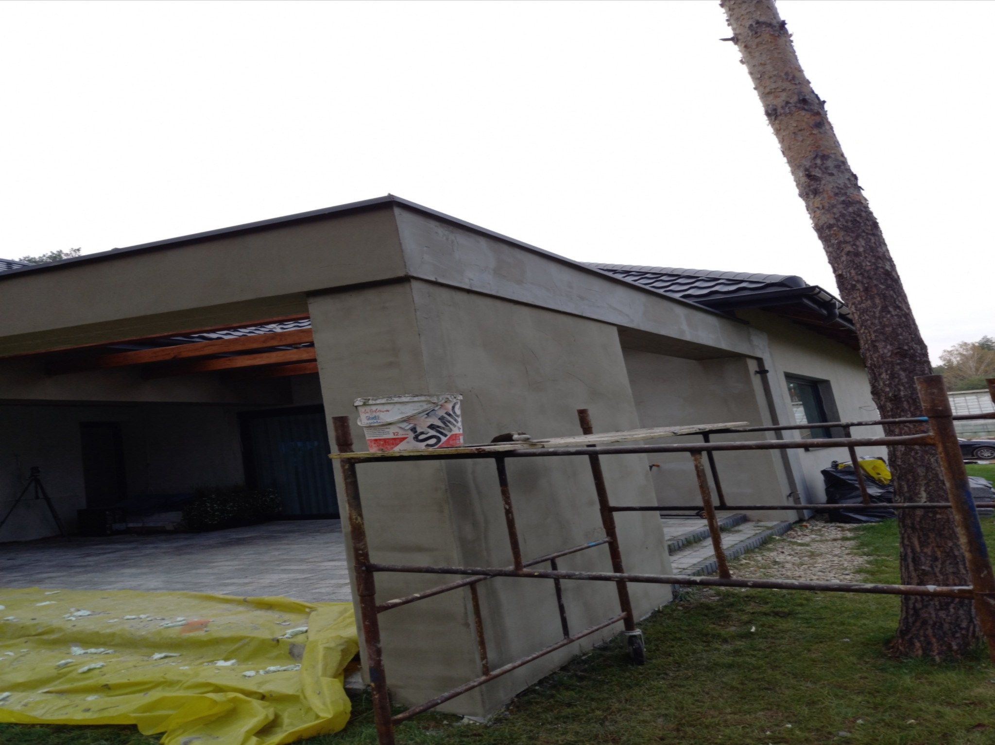 Elewacja budynku po nałożeniu świeżej warstwy szpachli, widoczne rusztowanie, wiadro z resztkami materiału oraz żółta folia ochronna na podłożu.