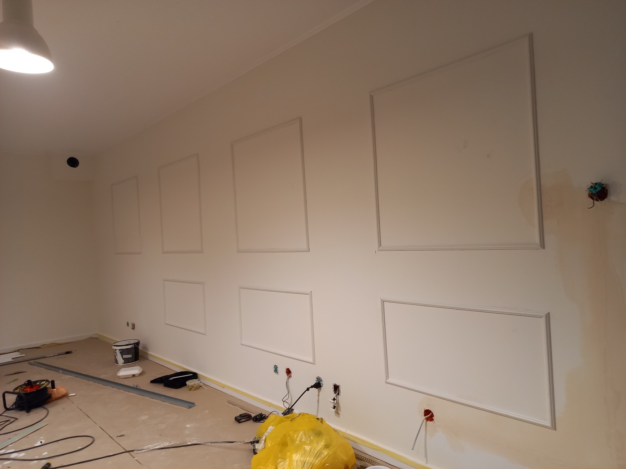 Ściana w trakcie dekorowania panelami ściennymi z listew, widoczne narzędzia, puszki elektryczne i zabezpieczona folią podłoga.