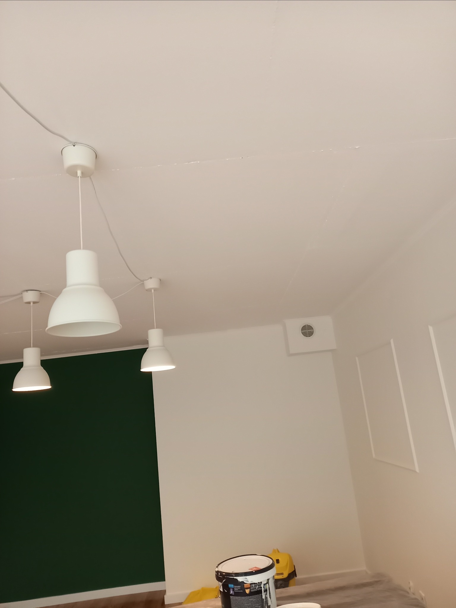 Wnętrze pokoju w trakcie malowania: ściana pomalowana na ciemnozielono, reszta ścian białe, widoczne trzy białe lampy sufitowe, wiadro z farbą i odkurzacz na podłodze zabezpieczonej folią.