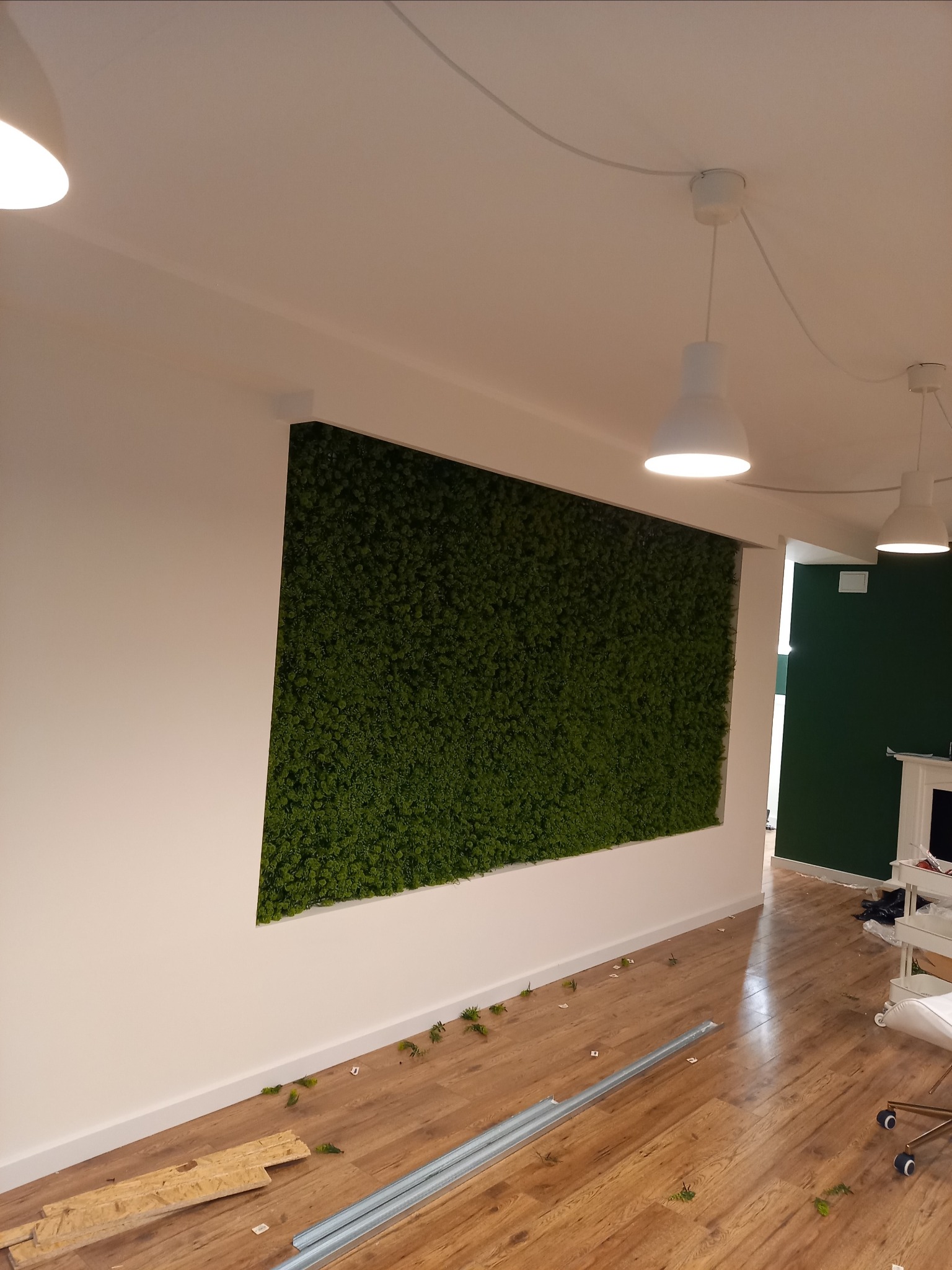 Prostokątny panel z mchu na białej ścianie w pomieszczeniu z drewnianą podłogą, widoczne elementy montażowe na podłodze, białe lampy wiszące i fragment zielonej ściany w tle.